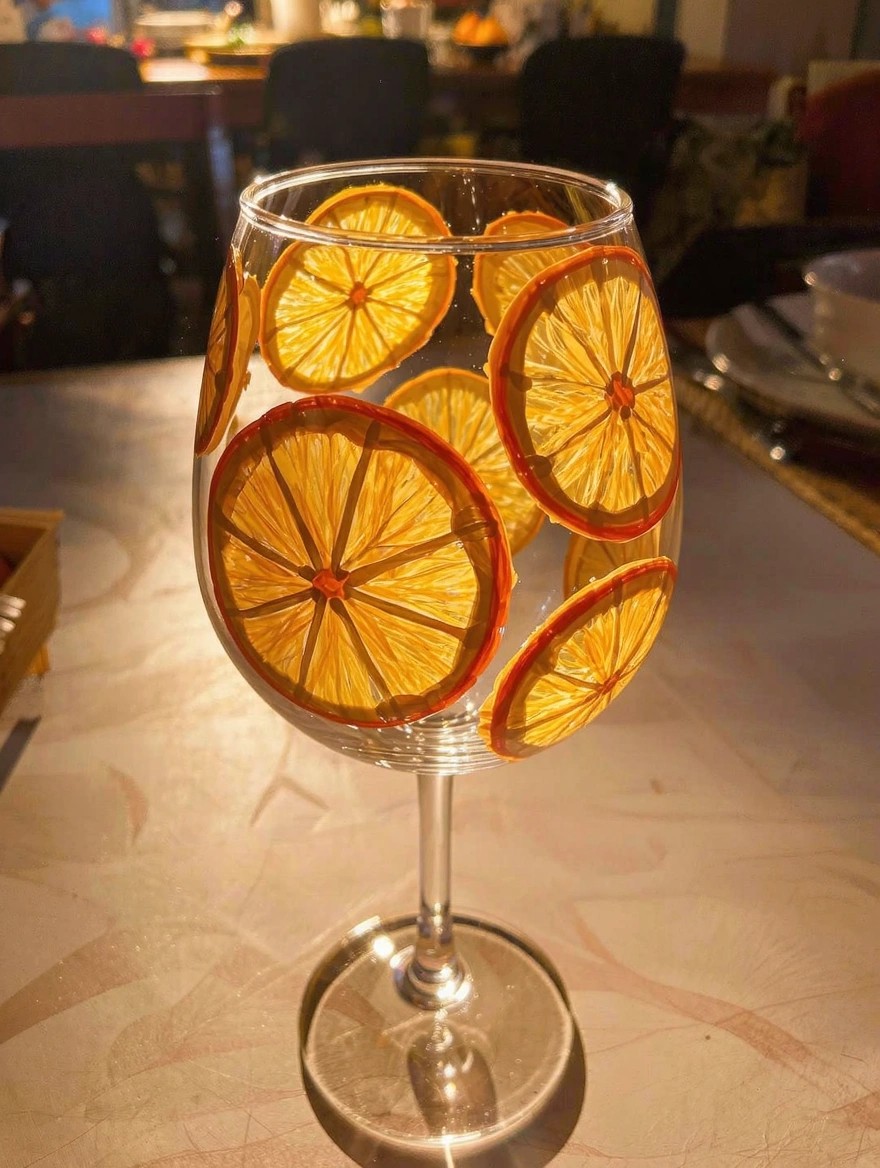 A clear stemmed wine glass painted with multiple overlapping orange slices in yellow and orange around the bowl sits on a table with warm lighting and reflections.