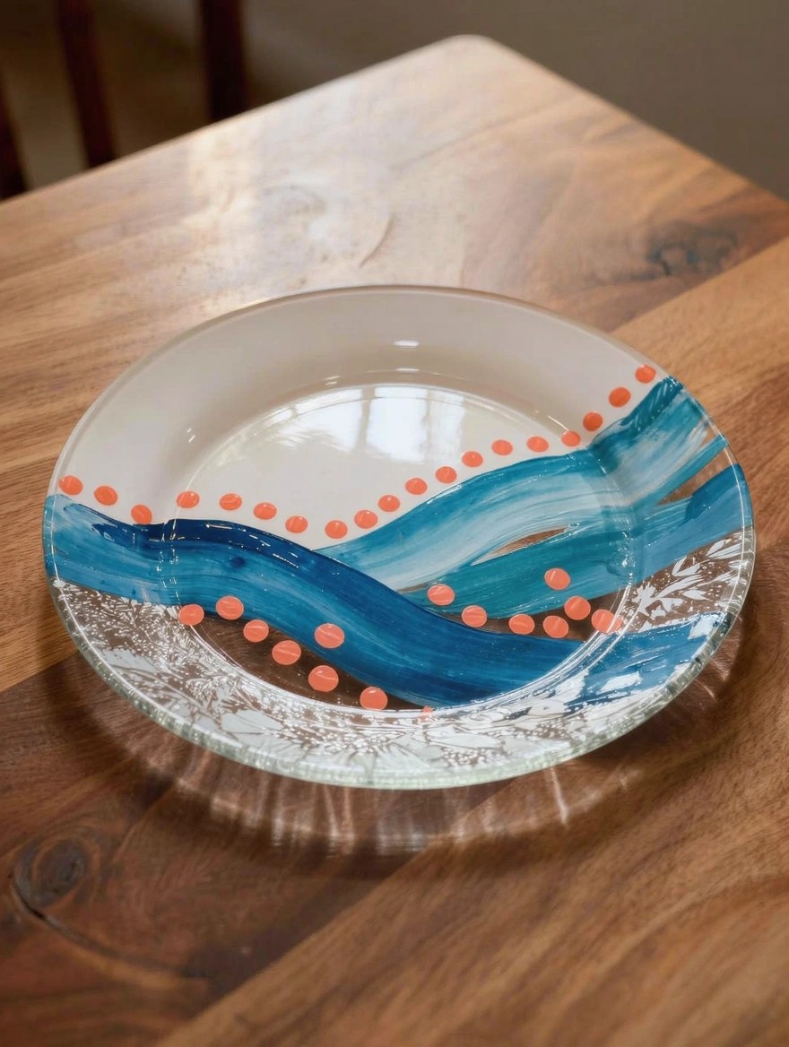 Clear round glass plate with hand-painted blue wavy lines and orange dots placed on a wooden table.
