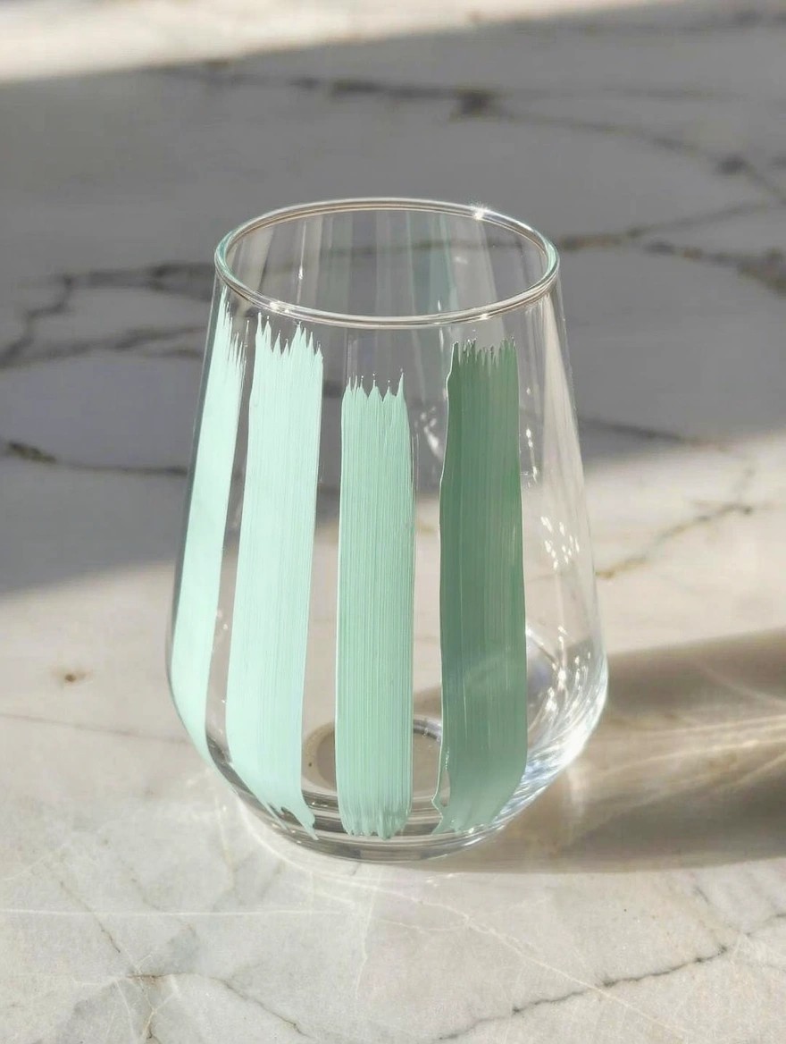 Clear glass tumbler with vertical mint green brushstroke stripes sits on a white marble surface in natural light.