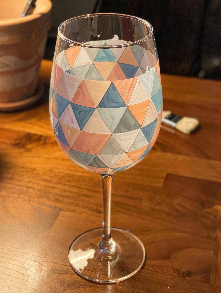 Stemmed wine glass painted with overlapping triangles in pastel peach, teal, gray, and white sits on a wooden table next to paintbrushes and a terracotta pot.
