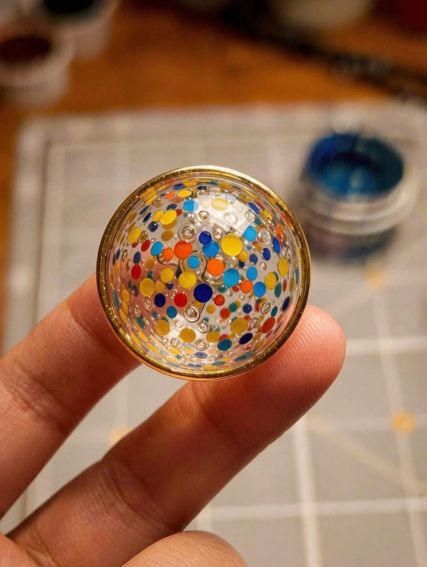 A hand holds a small clear glass dome with a gold rim, painted inside with multicolored polka dots, on a gray craft mat near paint jars.