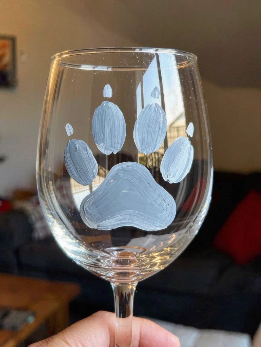 Clear wine glass with a large white paw print painted on the bowl, held by a hand against a home background.
