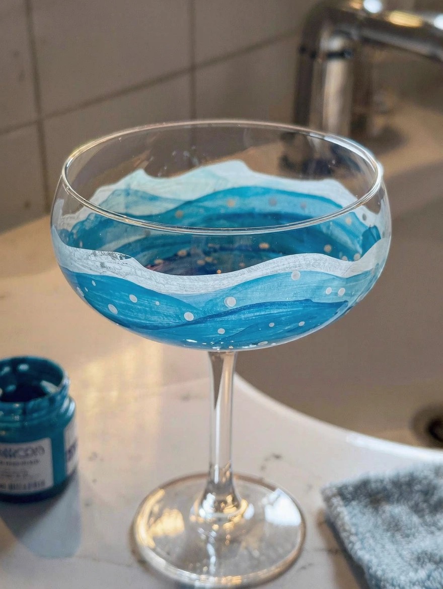 A coupe-style wine glass painted with blue ocean waves, white foam, and gold dots sits on a sink counter next to a jar of blue paint.