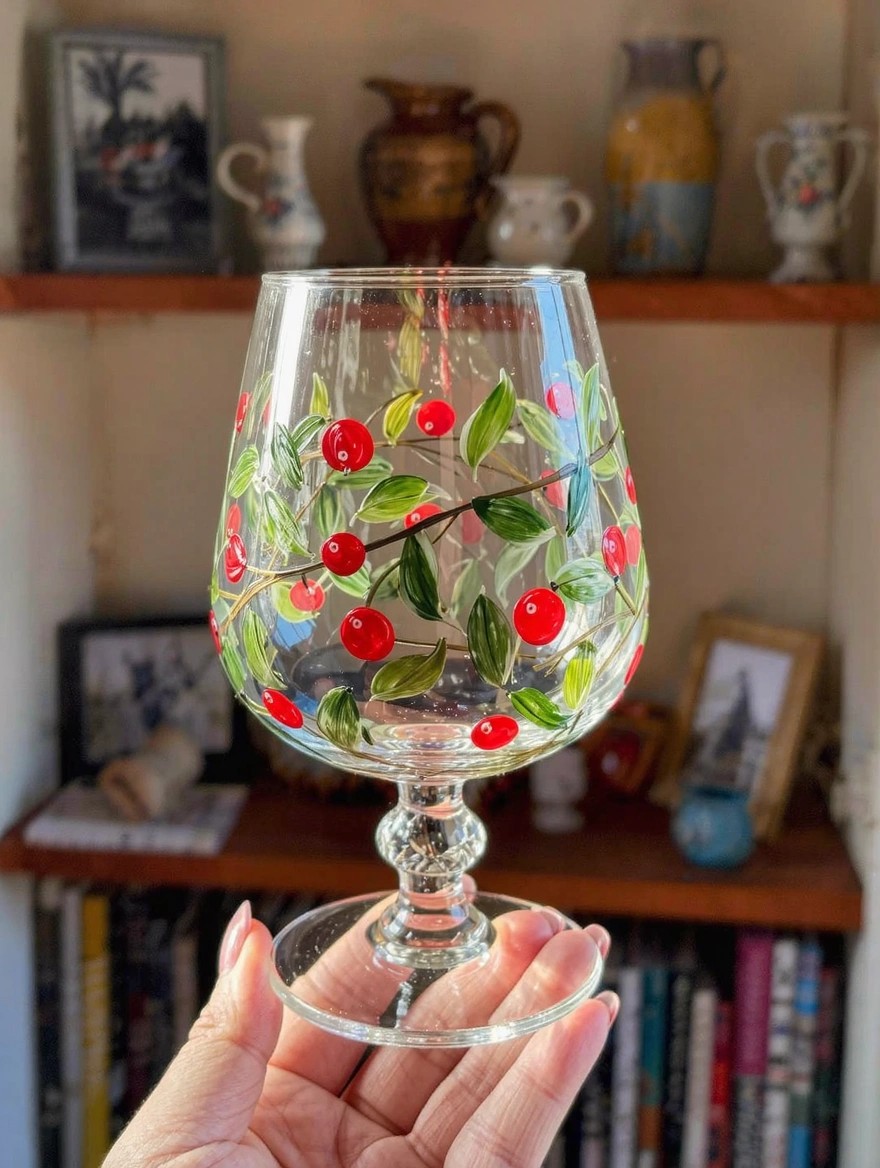 A hand holds a clear balloon wine glass hand-painted with red cherries and green leaves on a wooden shelf with ceramics and books in the background.