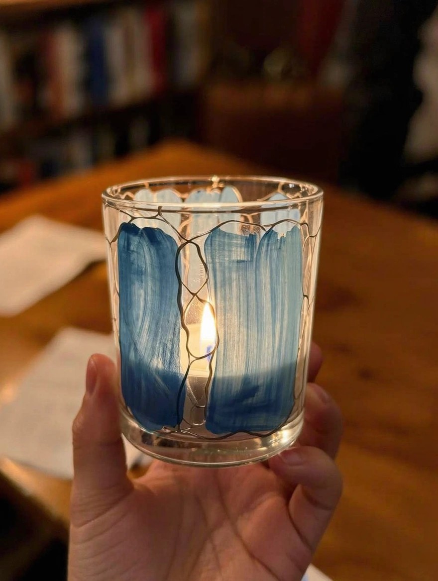 A hand holds a lit cylindrical glass candle holder painted with thick blue brushstrokes outlined in fine crackled lines, placed on a wooden table near bookshelves.