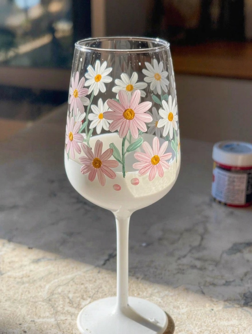 Clear stemmed wine glass hand-painted with clusters of white and pink daisies around the bowl, on a table next to a red paint tube.