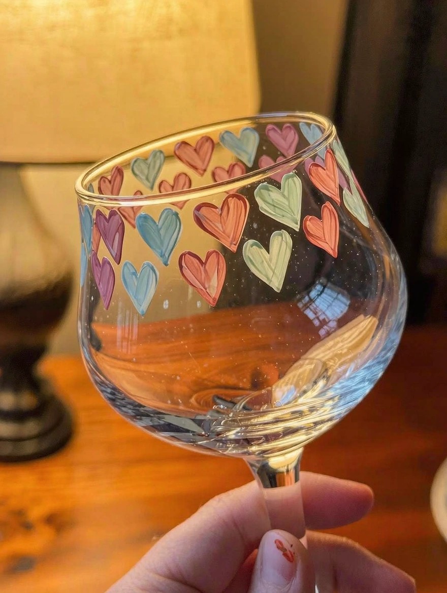 Clear tulip wine glass with hand-painted multicolored hearts clustered around the bowl, held by a hand on a wooden table near a lamp.