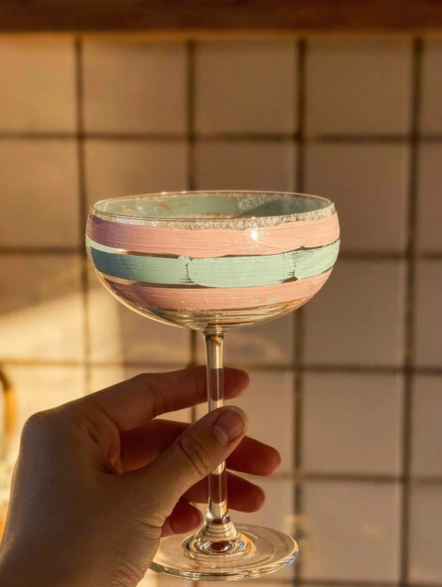 A hand holds a coupe wine glass with horizontal pink and teal painted stripes around the upper bowl against a green-tiled kitchen wall.