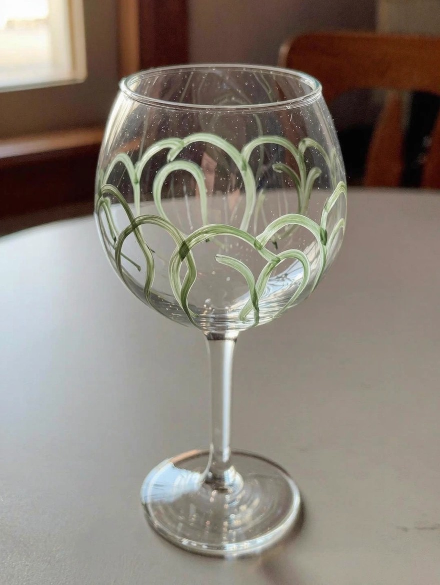 Clear stemmed wine glass with hand-painted green swirling vine-like scrolls on the bowl, placed on a table near a window.