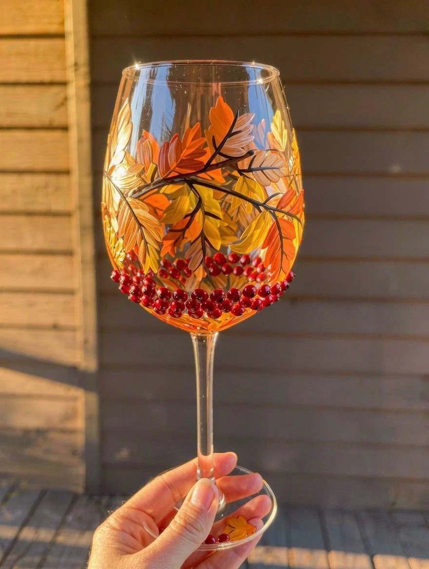 A hand holds a clear stemmed wine glass painted with orange, yellow, and gold autumn leaves around the bowl and red berries clustered at the base, set against a wooden outdoor background in golden light.
