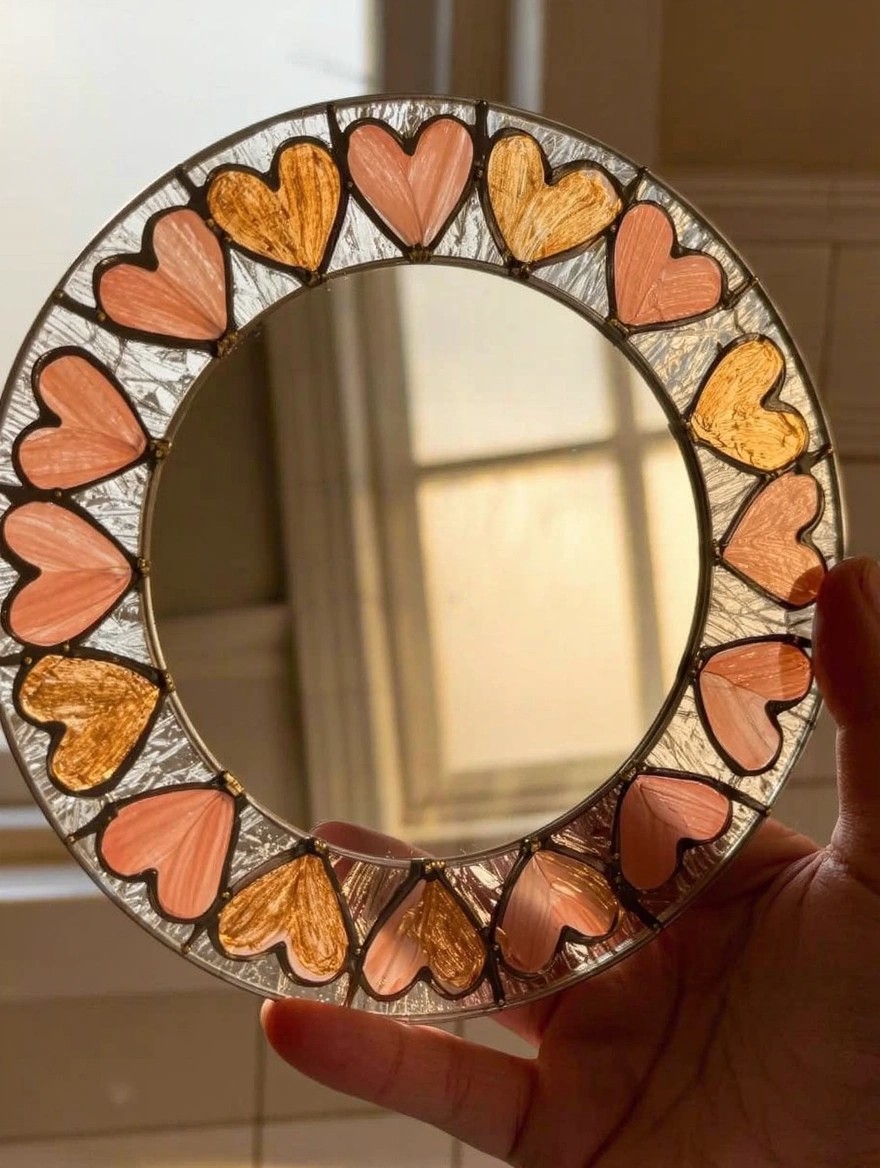 A hand holds a small round mirror framed by a stained glass-style border of alternating pink and orange painted hearts with black leading lines.