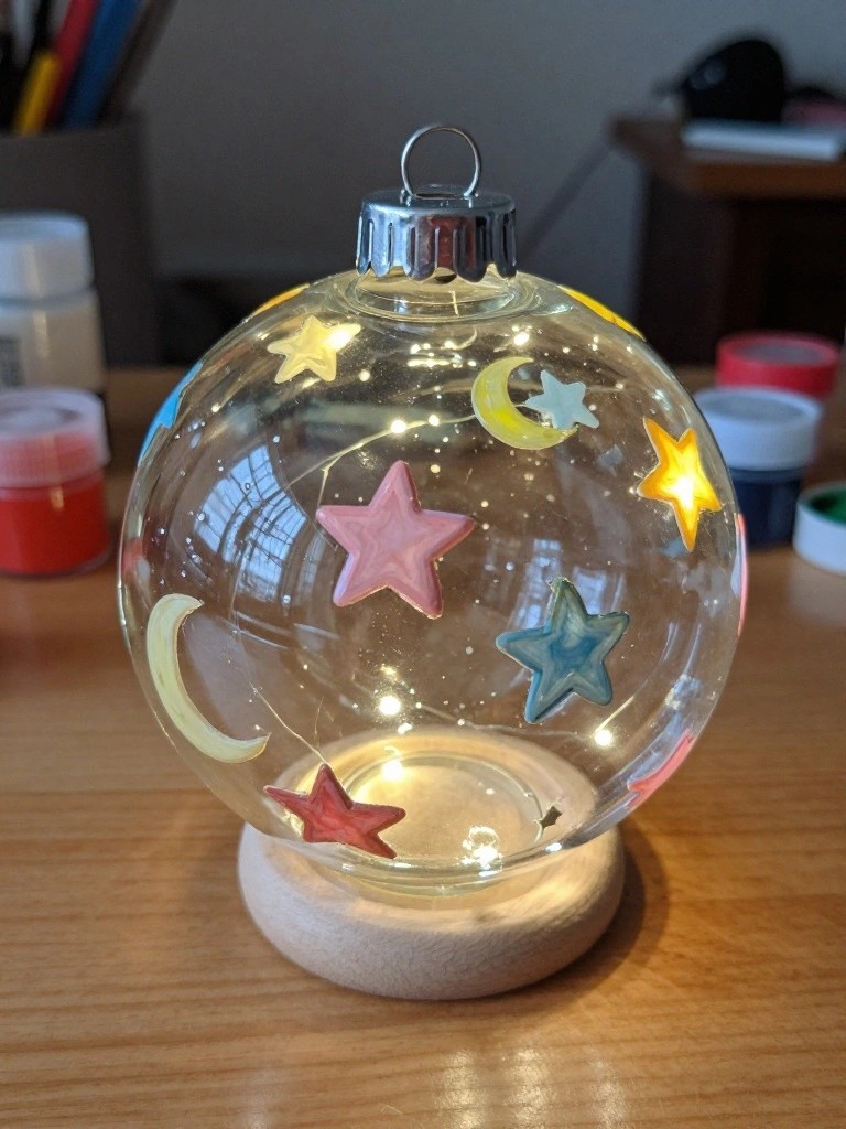 A clear glass sphere ornament painted with stars and moons in pink, blue, yellow, and red sits on a wooden base with string lights inside.
