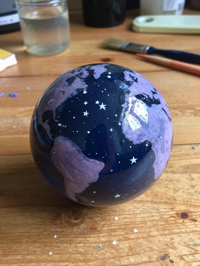 Painted blue sphere with purple continents, stars on wooden table amid art supplies