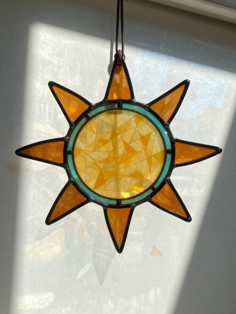 Stained glass sun with orange rays, yellow geometric center, and teal border hanging in sunlight.