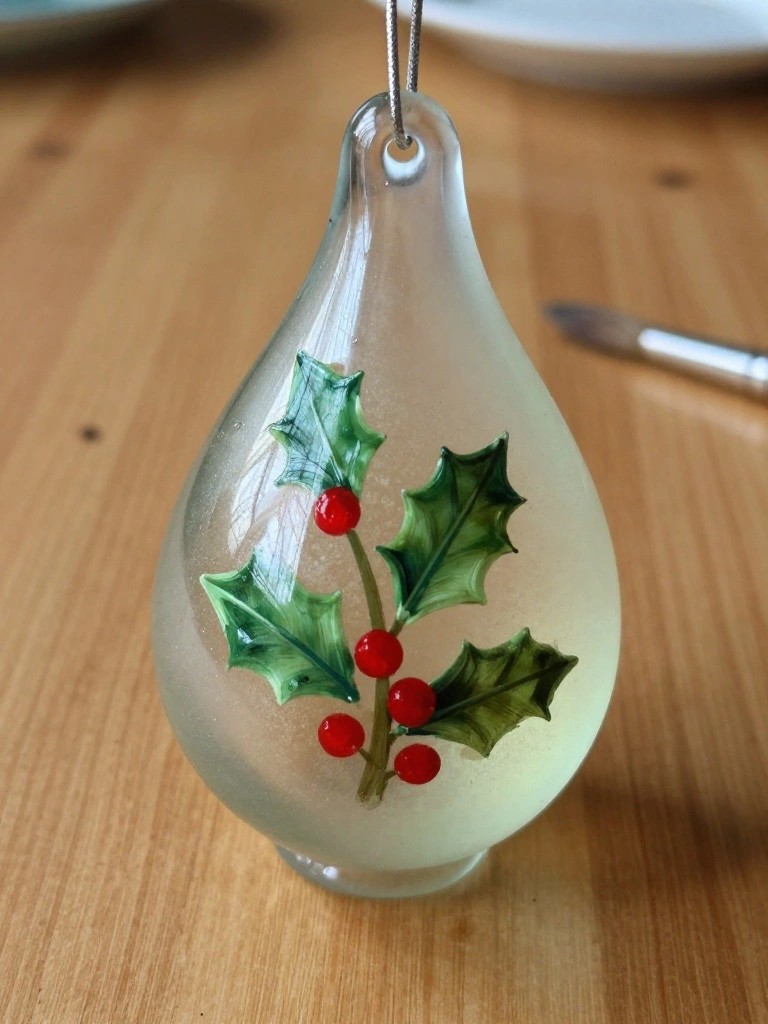 Glass teardrop ornament with green holly leaves and red berries on wood