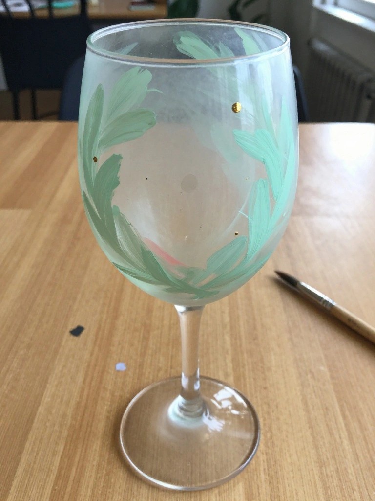 Wine glass hand-painted with green leaves and gold dots on wooden table beside paintbrush