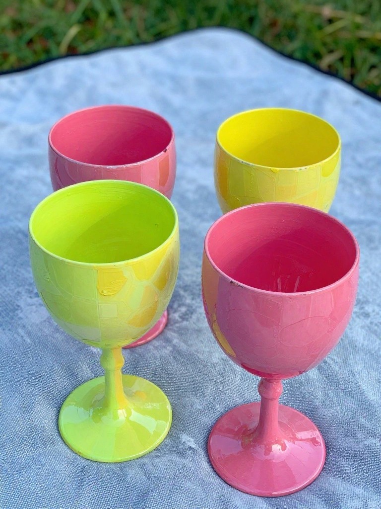 Four bright hand-painted goblets in pink, yellow, and green on blue blanket.