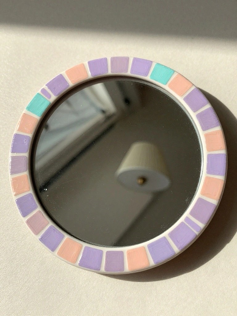 A small round mirror with a white frame edged in hand-painted squares of pastel purple, peach, and teal arranged like mosaic tiles.