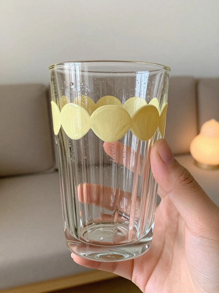 A hand holds a clear ribbed glass cup with a yellow scalloped painted border near the top.