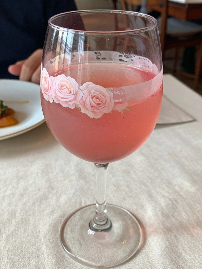 Wine glass of pink liquid adorned with pink roses on white tablecloth.