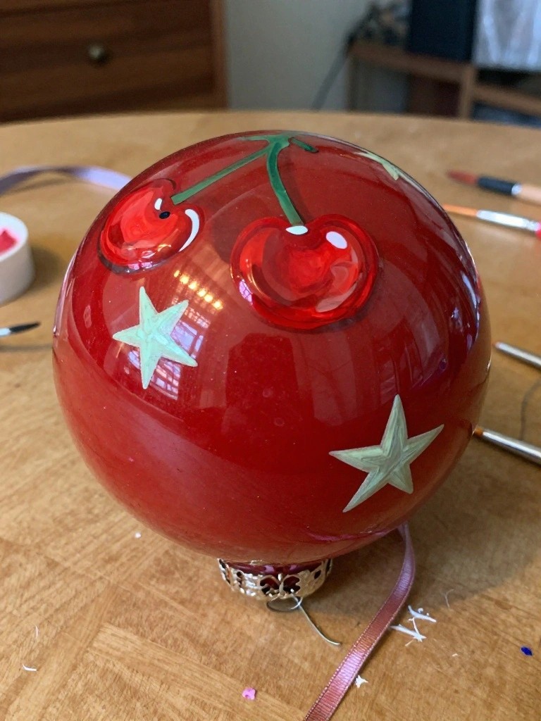 Shiny red ornament with cherries and stars on wooden table amid paint supplies