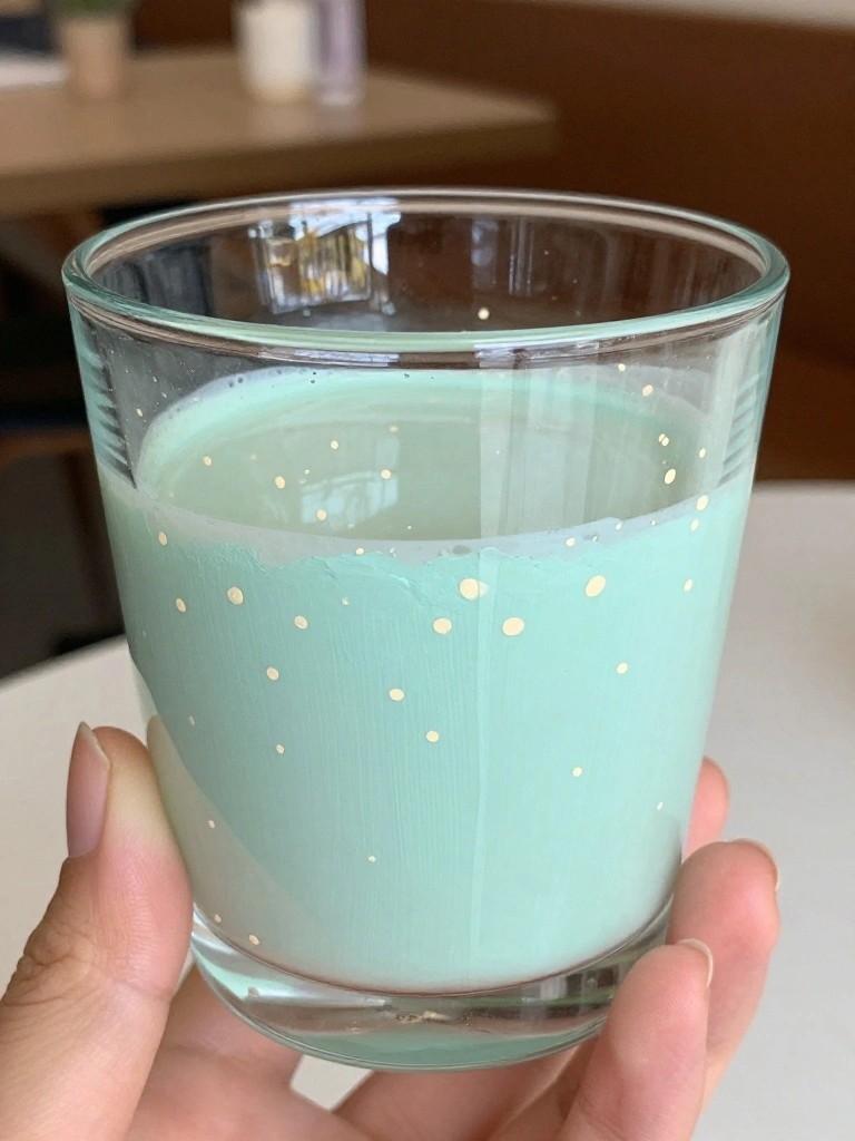 Hand holding glass jar candle with mint green wax and gold dots.
