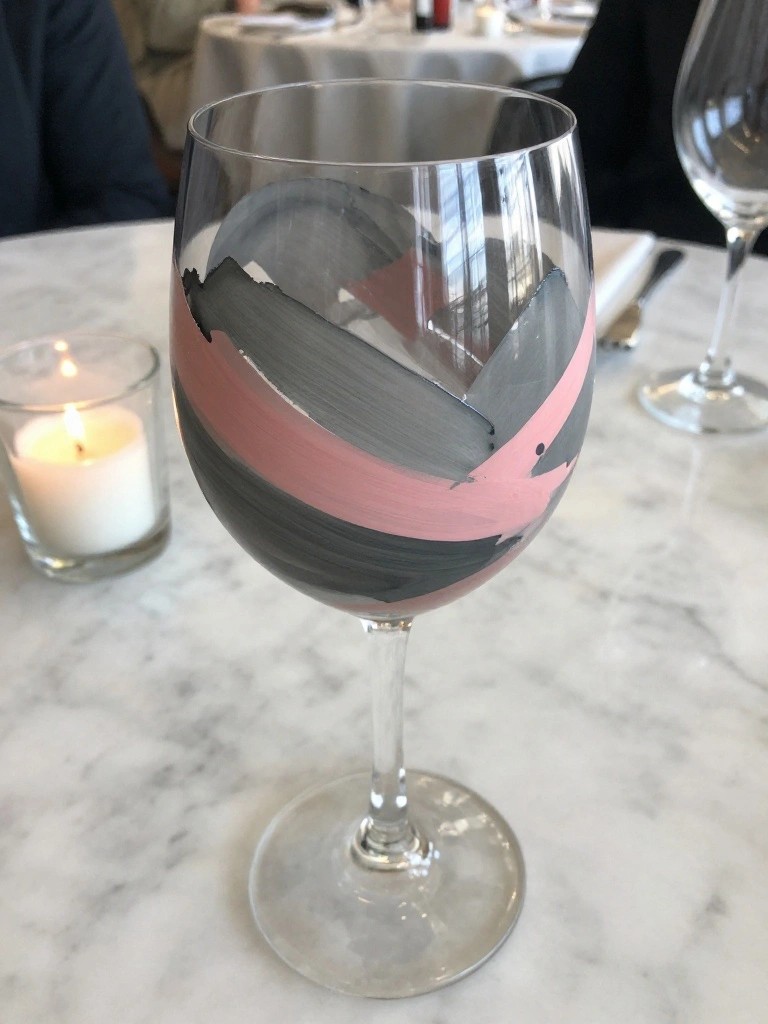 Wine glass with wide pink and gray brush strokes painted around the bowl on a white marble table.