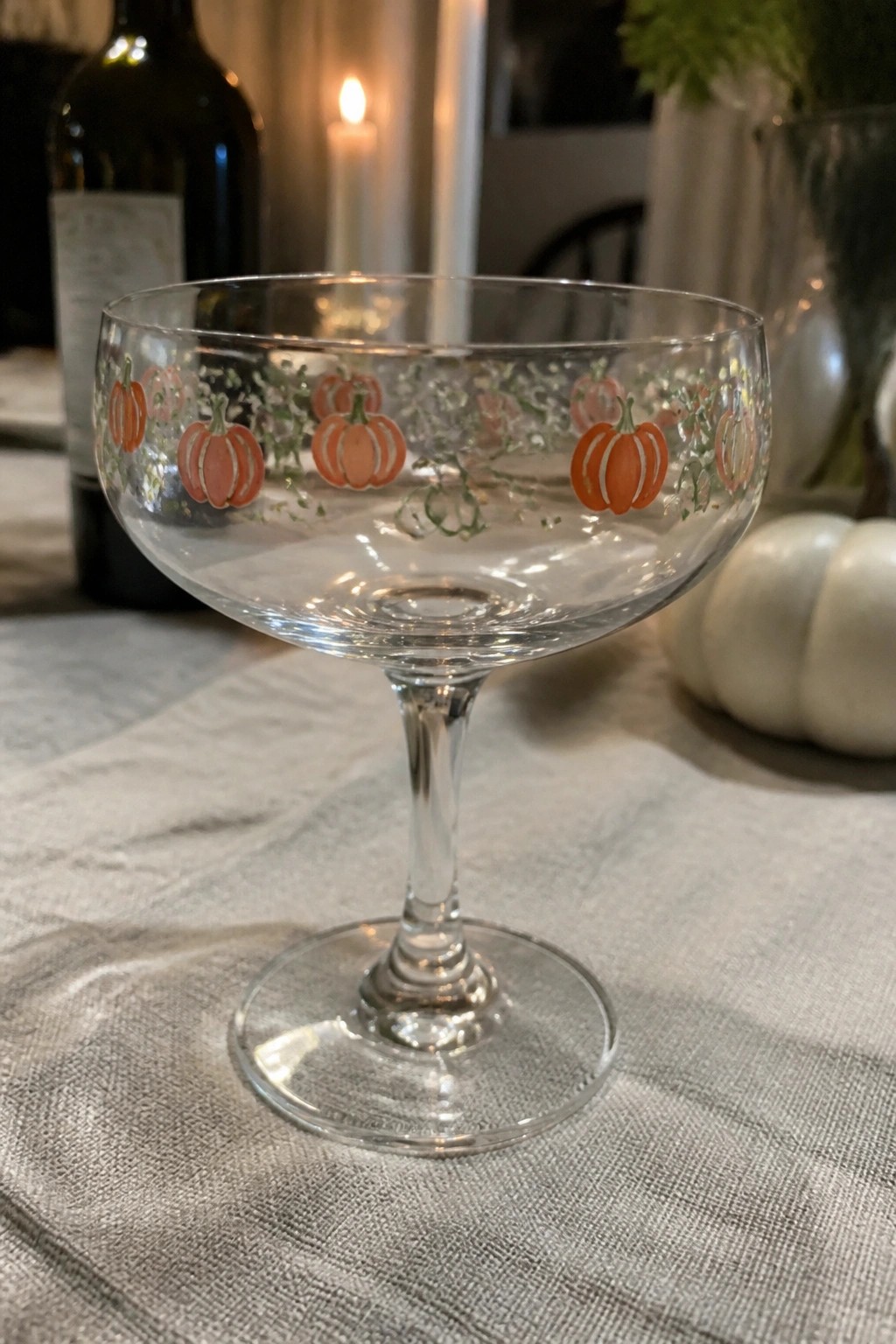 Clear coupe glass with orange pumpkin motifs and green vines on gray linen.