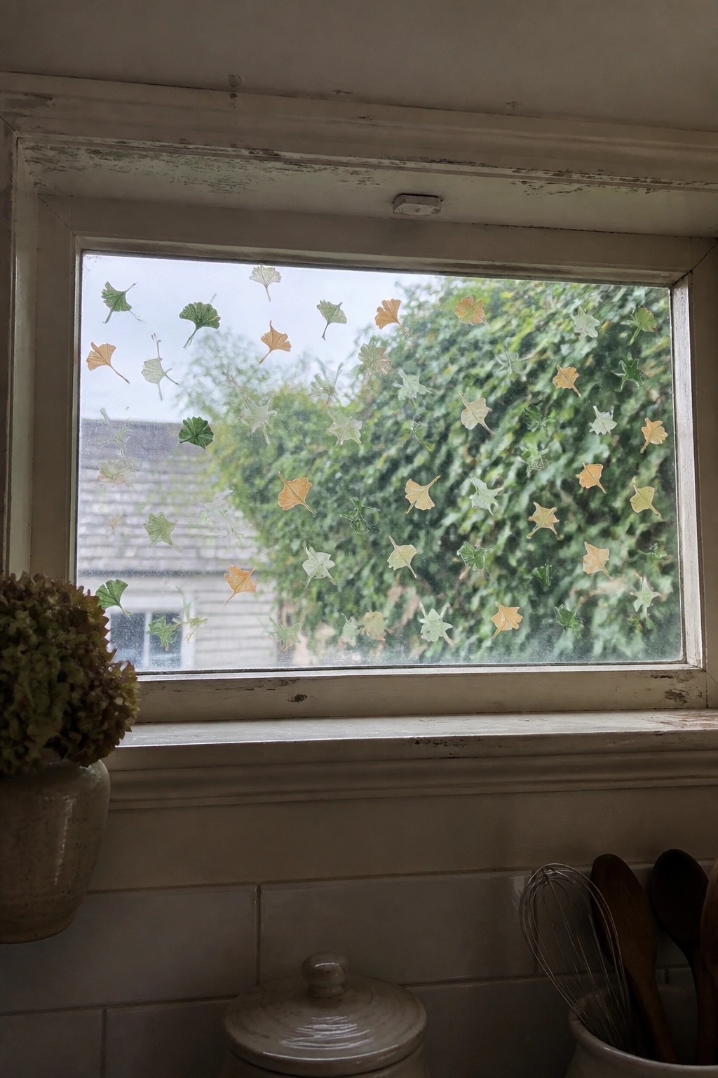 Window with colorful leaf decals overlooking greenery, utensils and vase on white sill