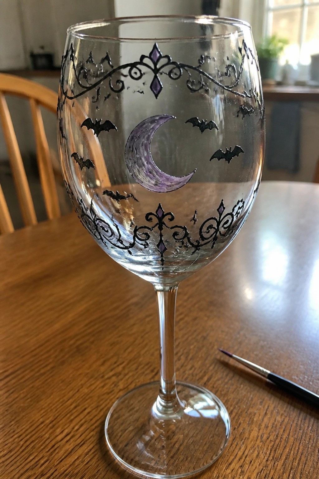 A clear wine glass with black ornate borders, a purple crescent moon, and black bats painted on the bowl rests on a wooden table next to a paintbrush.