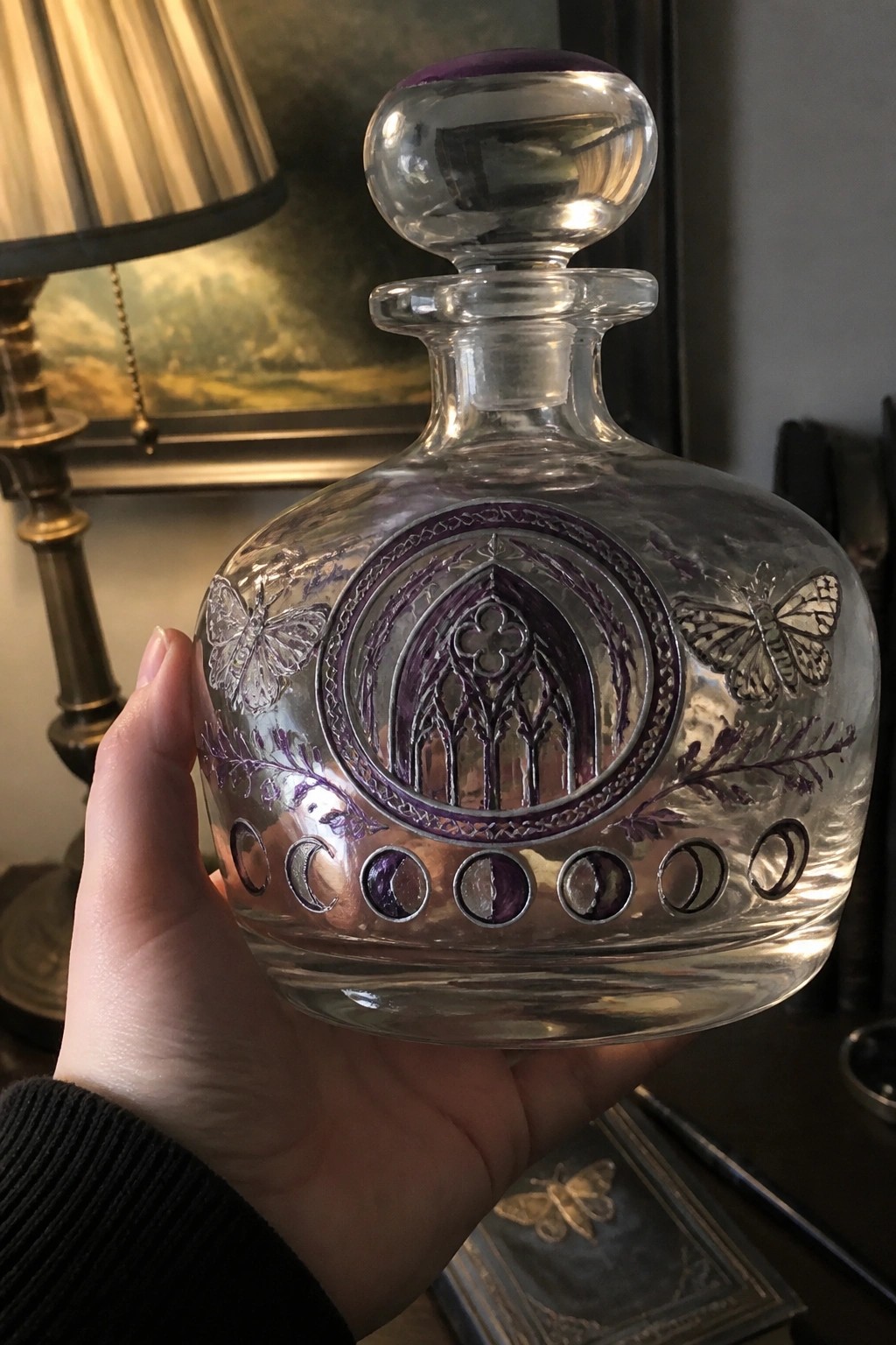 Hand holding clear glass decanter with purple moon phases, butterflies, and gothic engravings