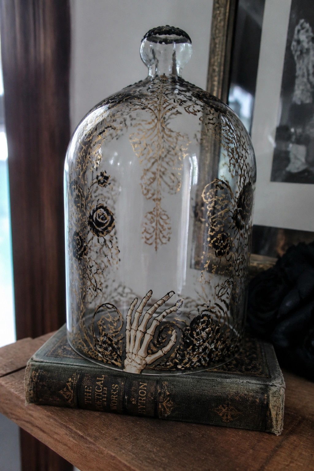 Glass cloche with gold-black roses and skeletal hand atop Byron poetry book.