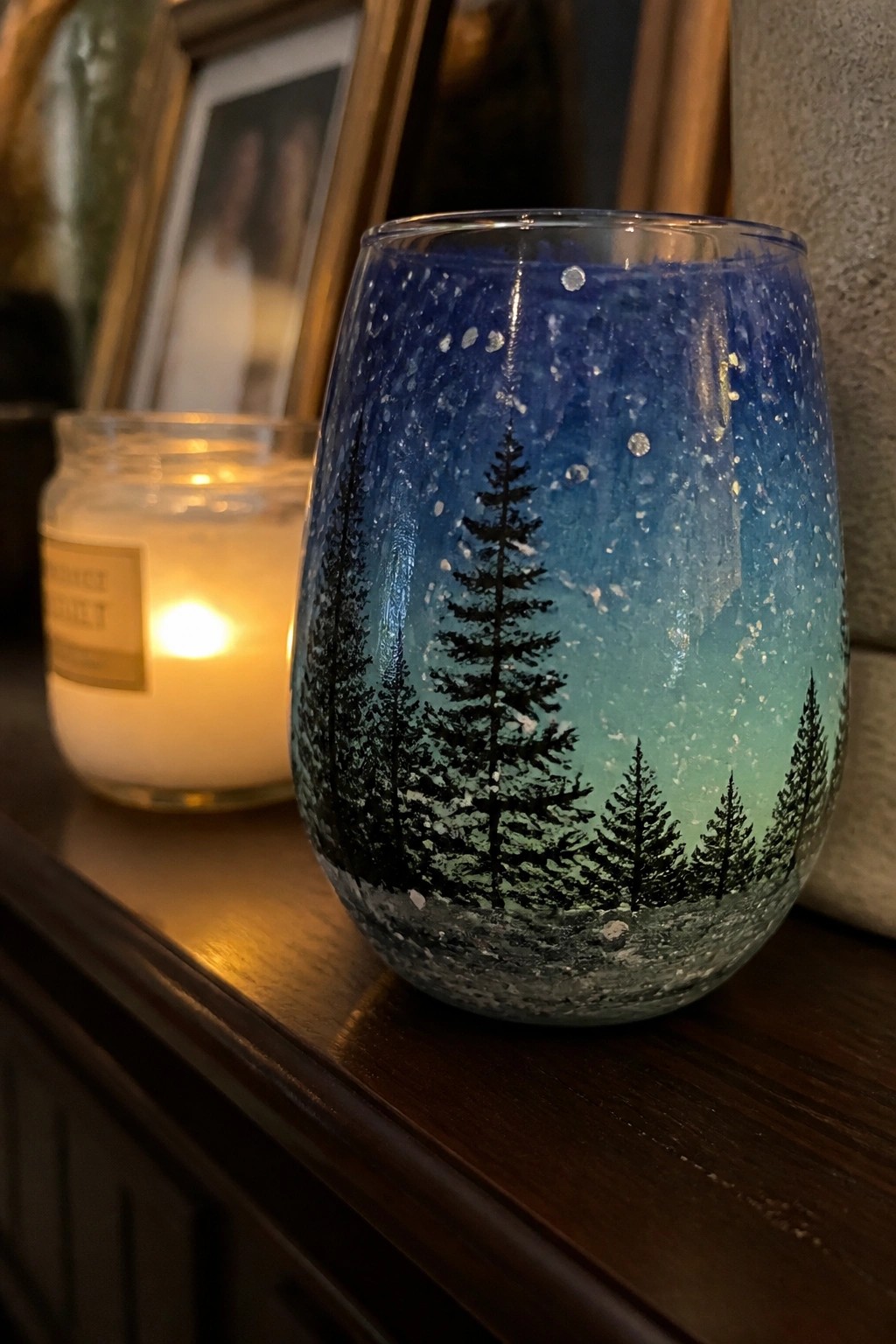 Blue painted glass with pine tree silhouettes under starry sky beside lit candle on wood