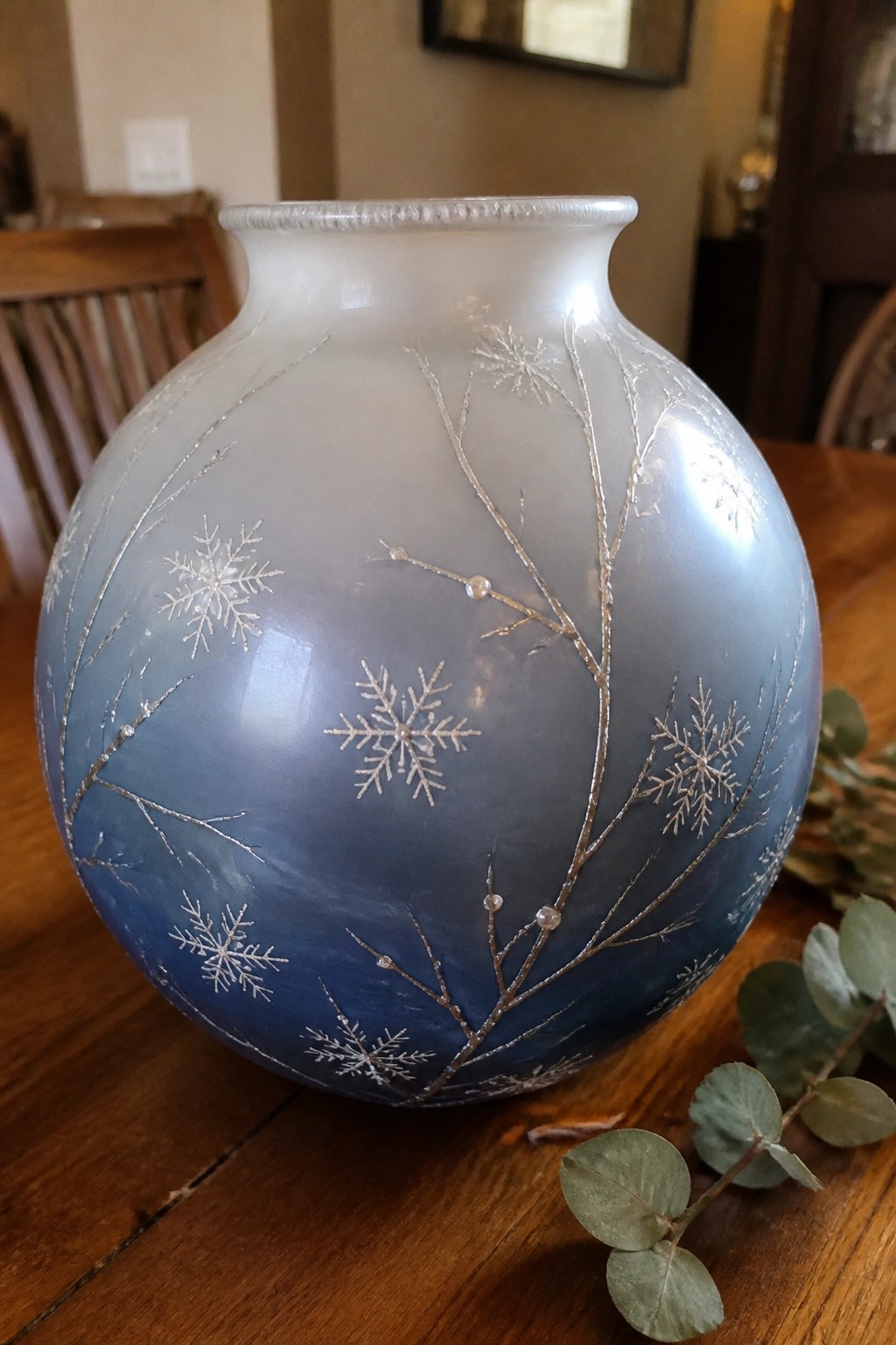 A round glass vase with blue gradient and silver snowflake and branch designs sits on a wooden table.