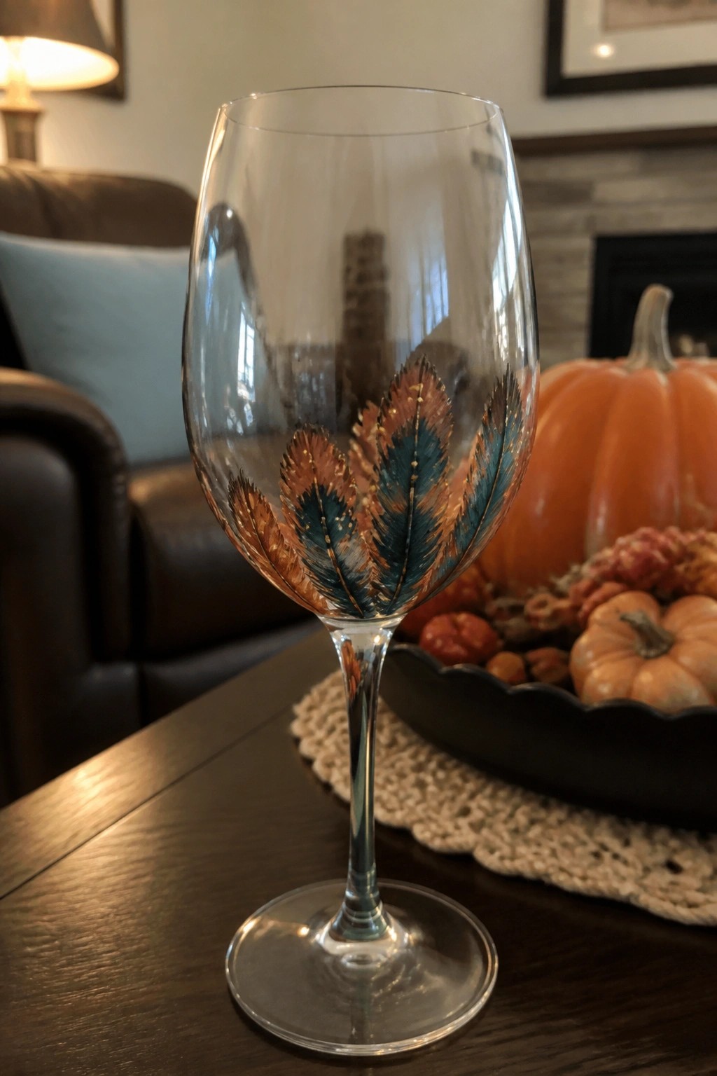 Clear wine glass with teal, brown, and gold feather paintings on a table beside a black tray of small pumpkins.