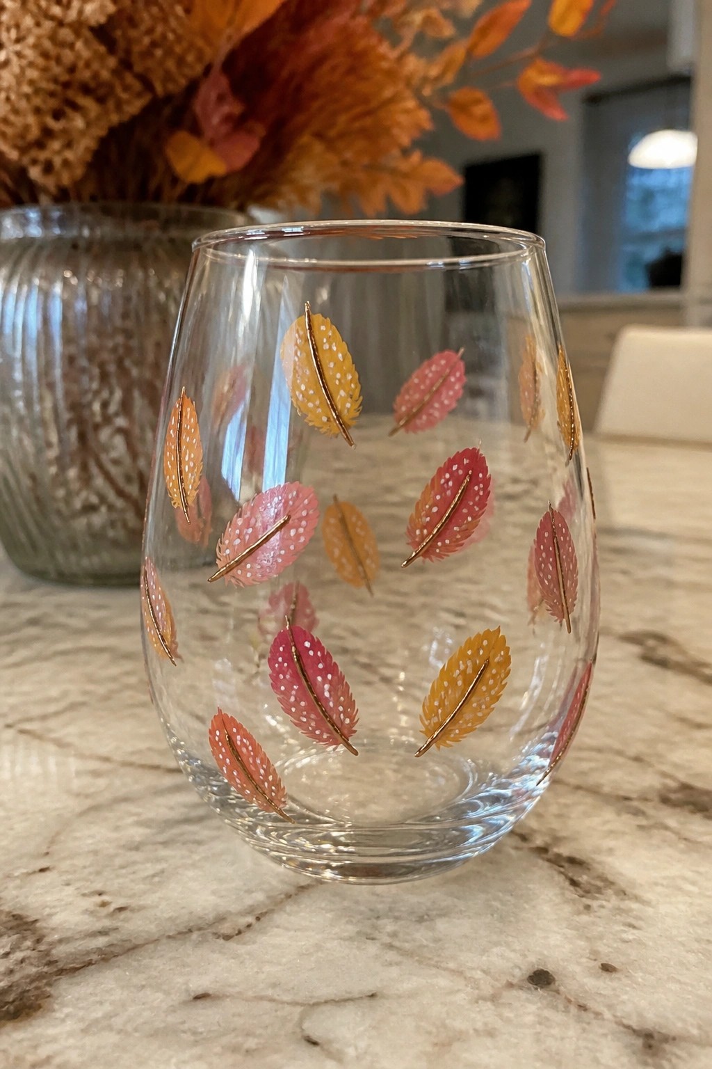 Stemless glass tumbler with colorful autumn feather designs on marble countertop.