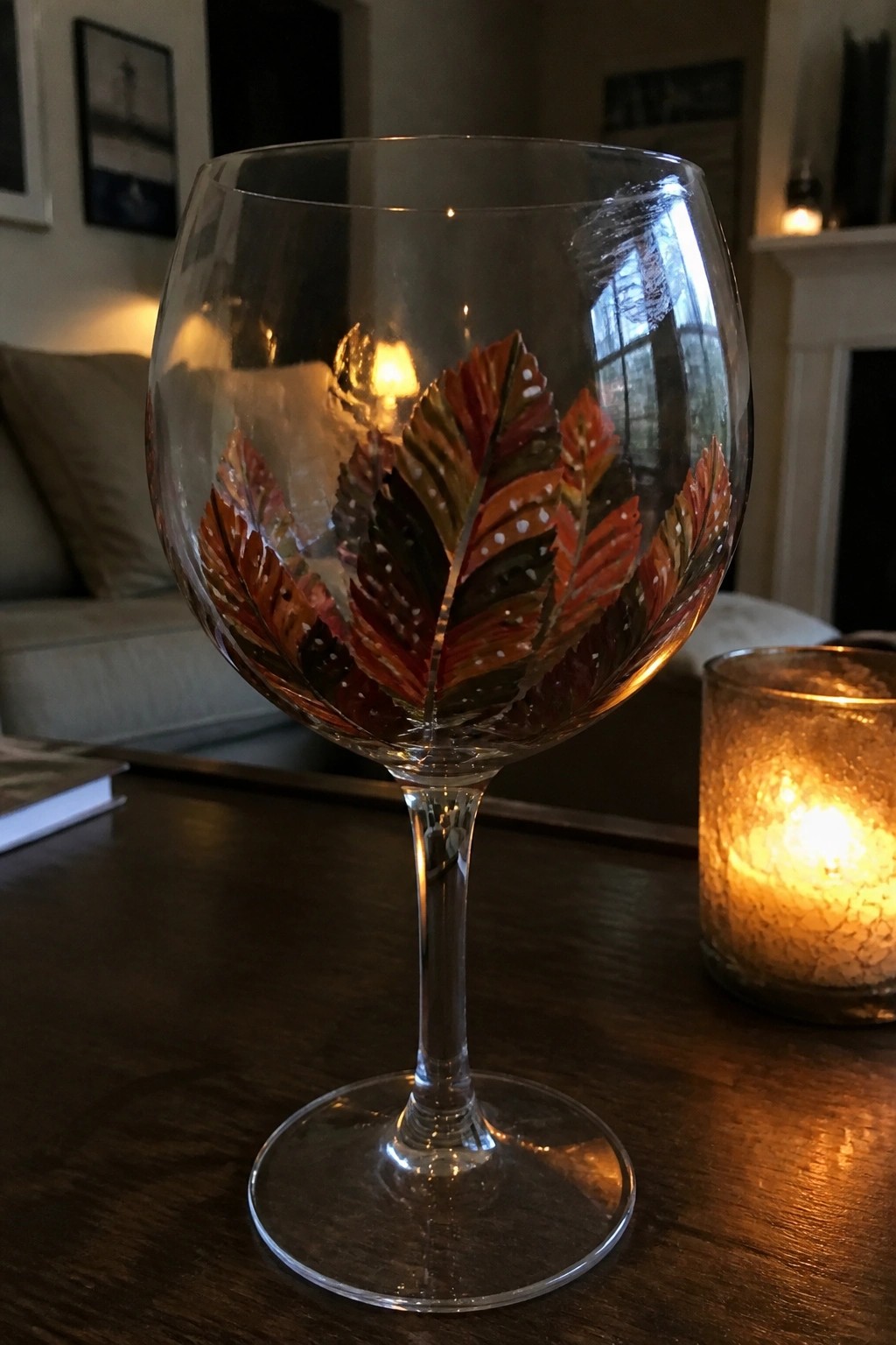 Painted wine glass with red and orange autumn leaves beside lit candle on table