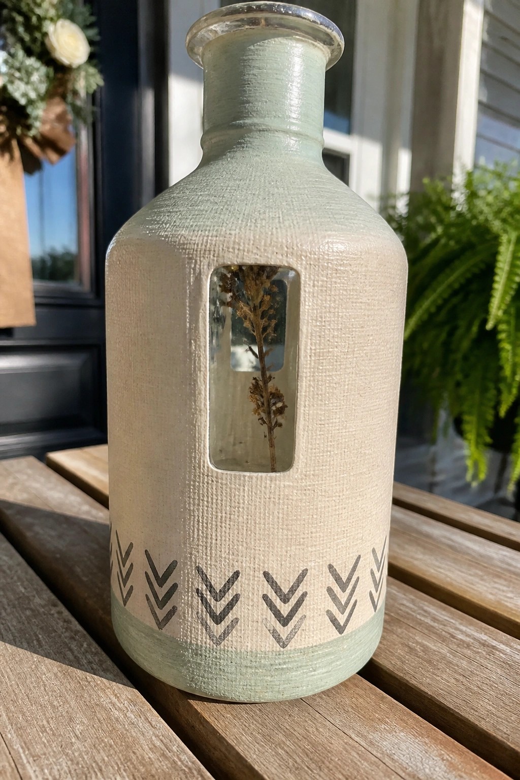 Painted glass bottle with rectangular window showing dried flowers and dark chevron border near the base on a wooden surface.