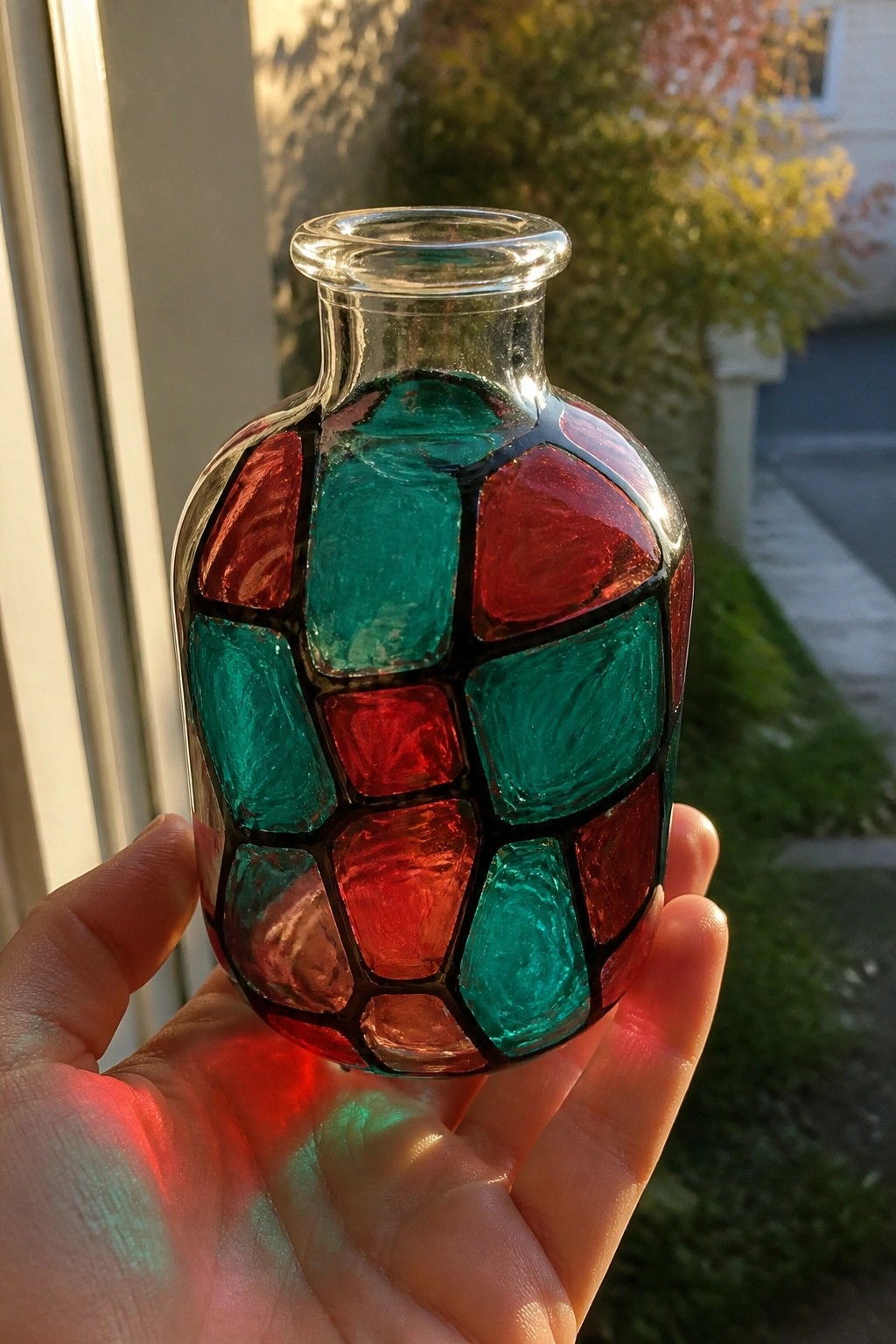 Hand holding sunlit glass bottle with red and teal mosaic patterns.