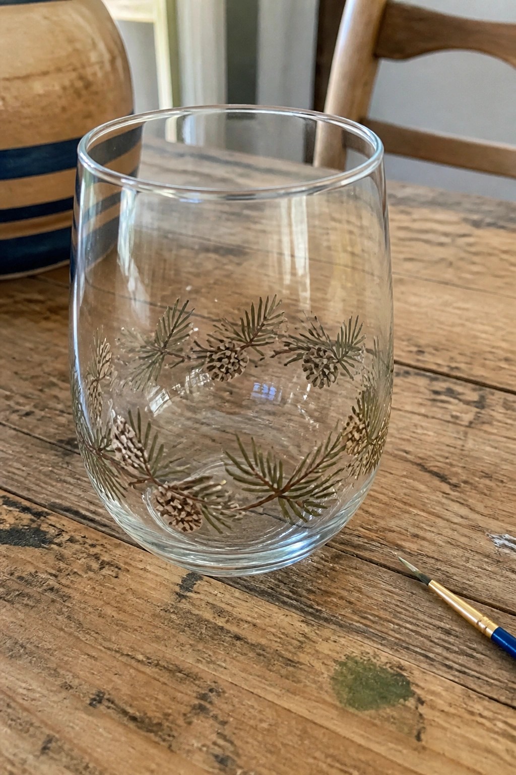 Clear glass tumbler hand-painted with pine branches and cones beside paintbrush on wood table