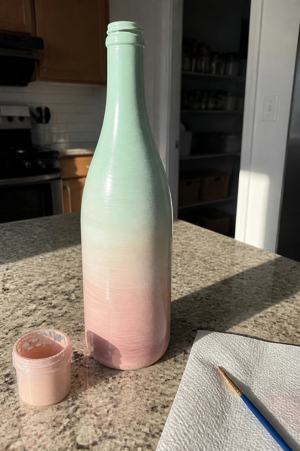 Ombre mint green to pink painted bottle with paint jar and brush on kitchen counter