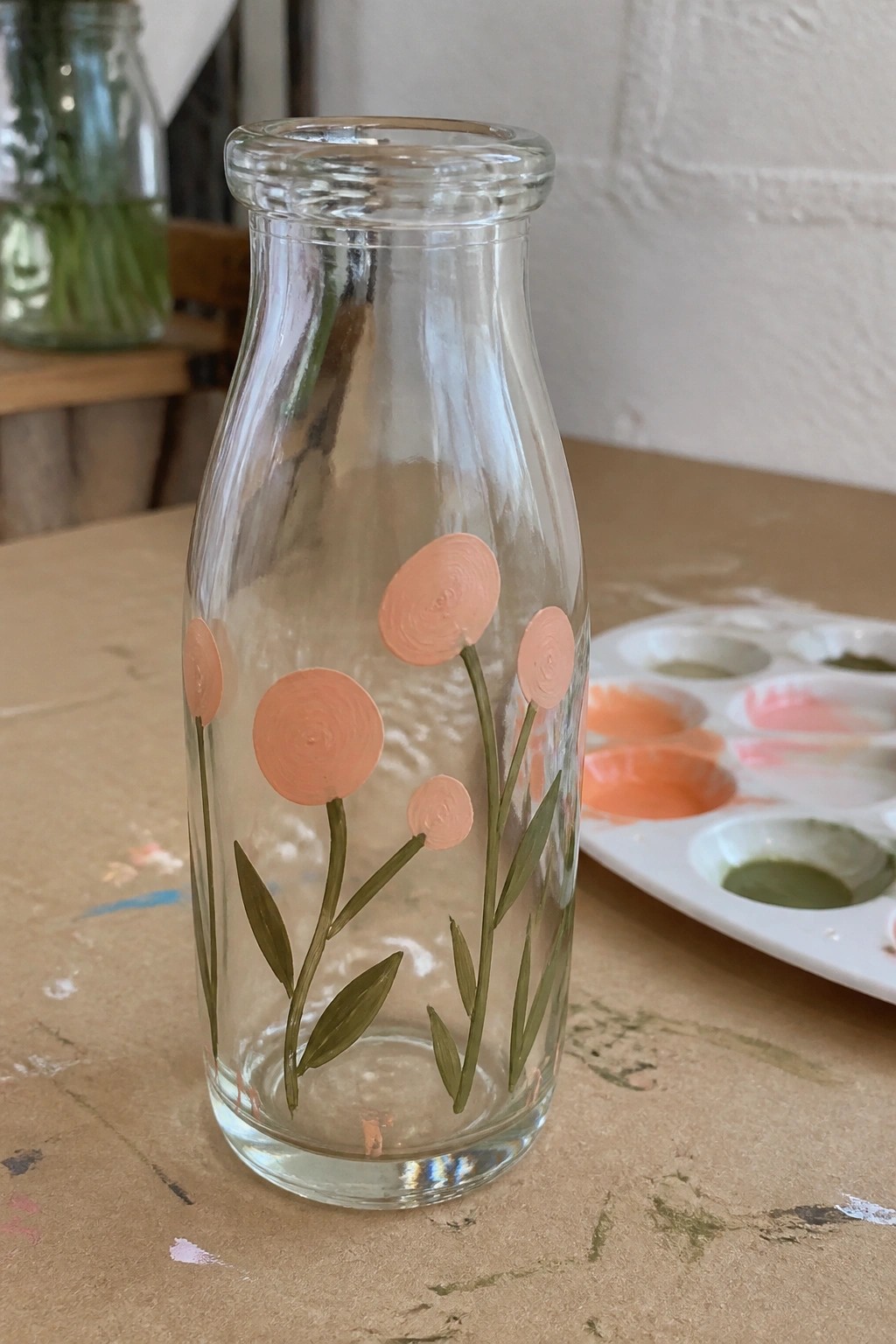 Clear glass bottle hand-painted with peach flowers and green stems beside paint palette