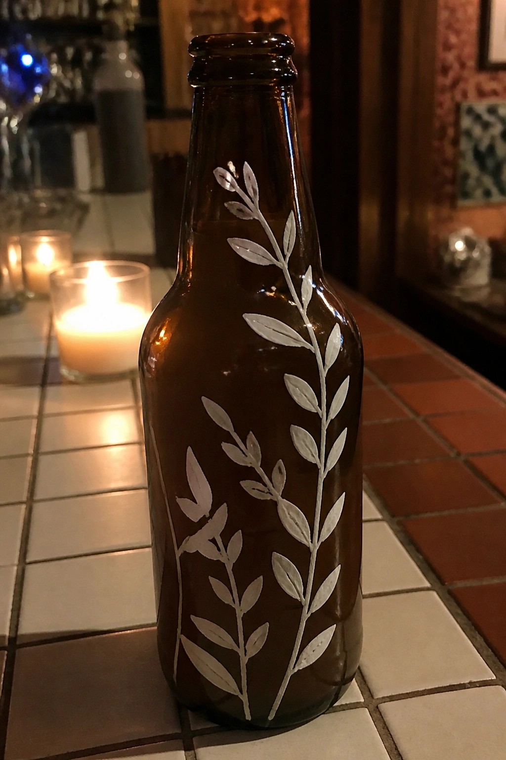 Brown glass bottle with white leaf etchings on tiled counter near lit candles