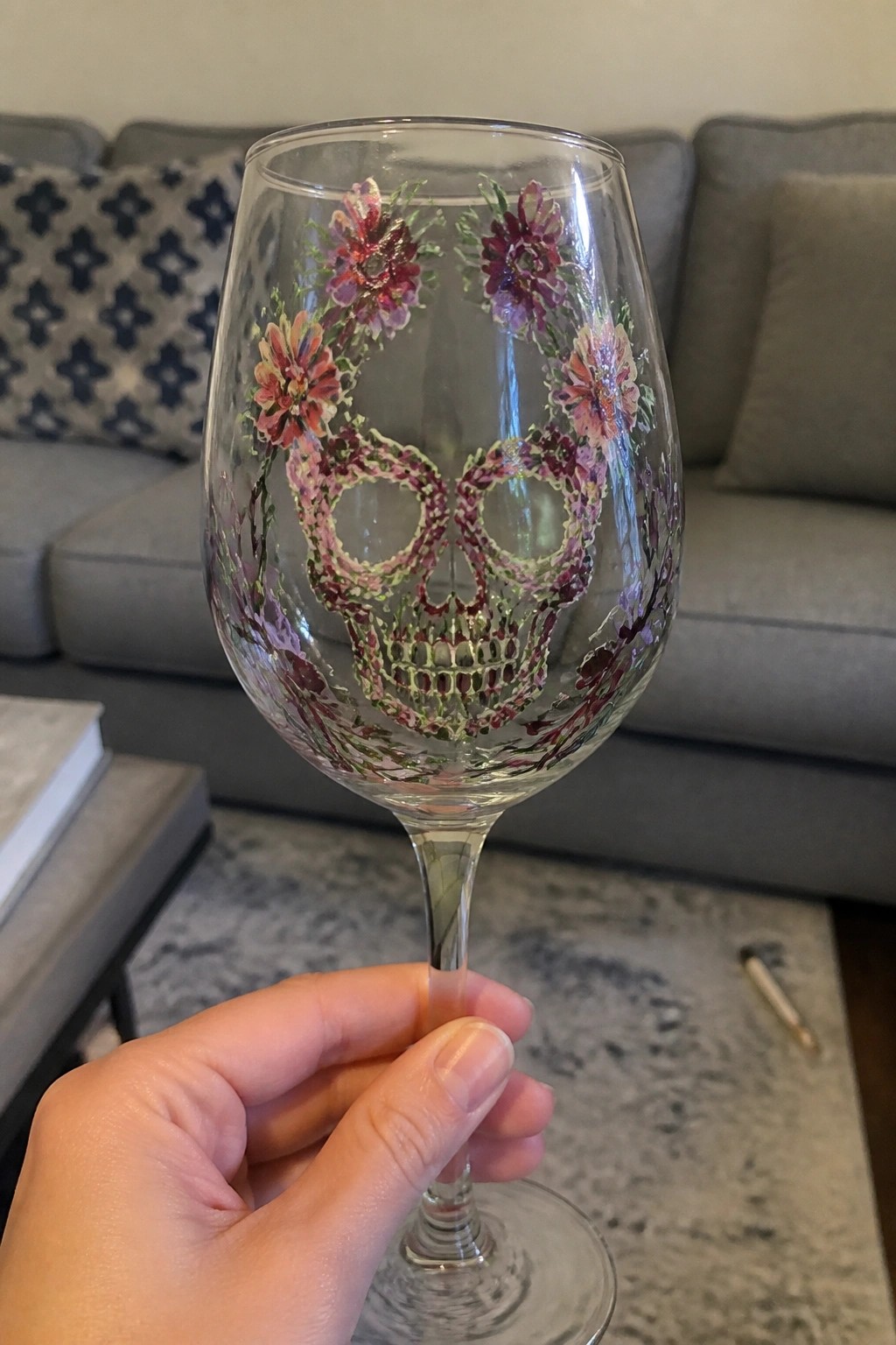 Hand holding wine glass with colorful floral skull painting in pink and purple.