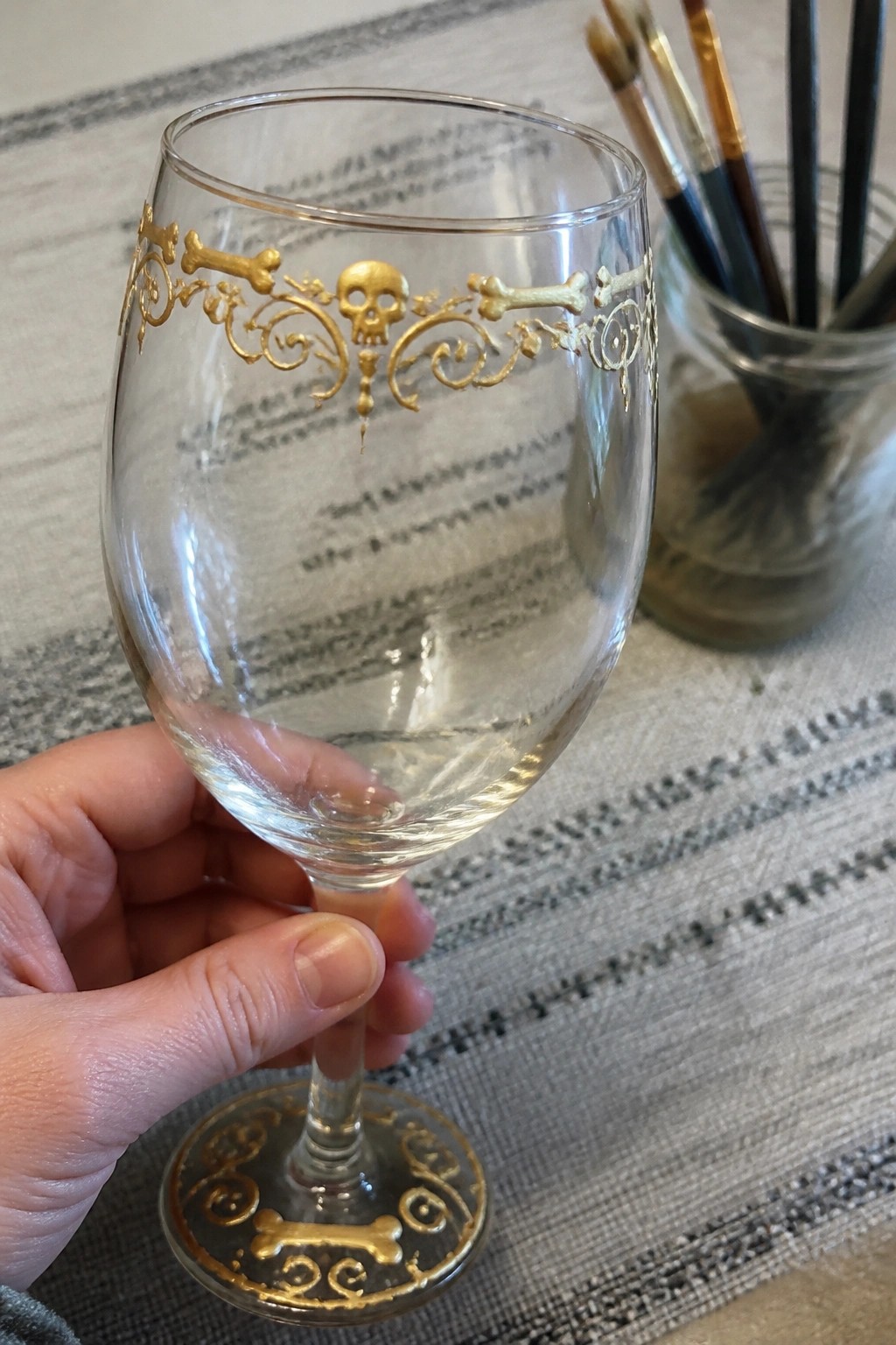Hand holding clear wine glass with gold skull, bones, and filigree designs.