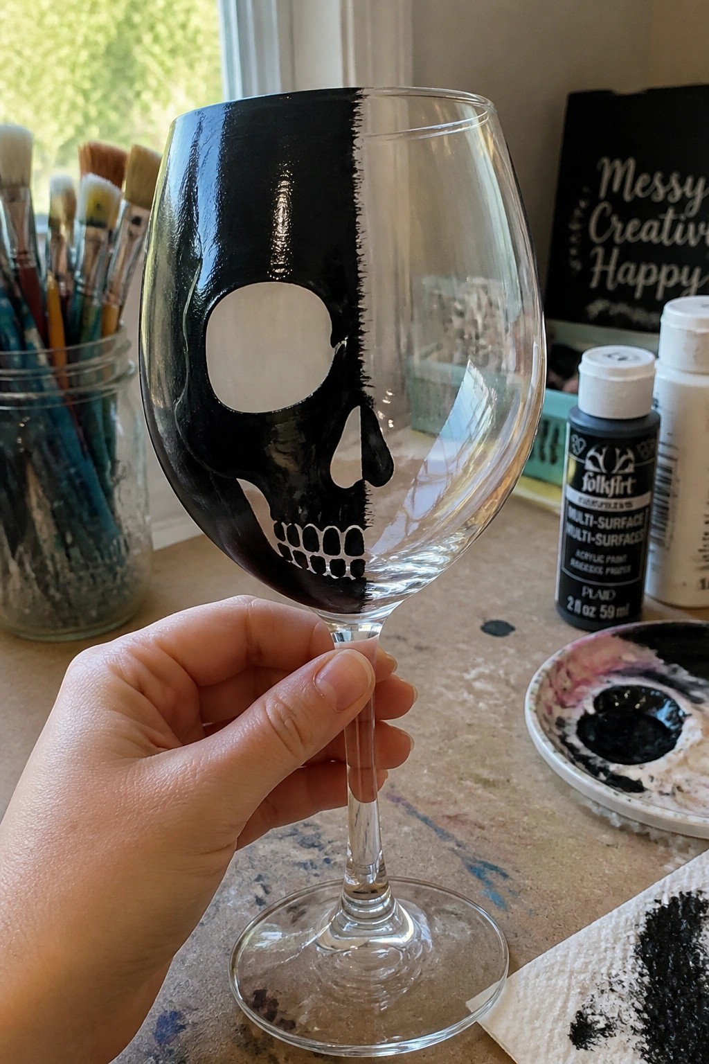 Hand holding wine glass with black skull design amid art supplies