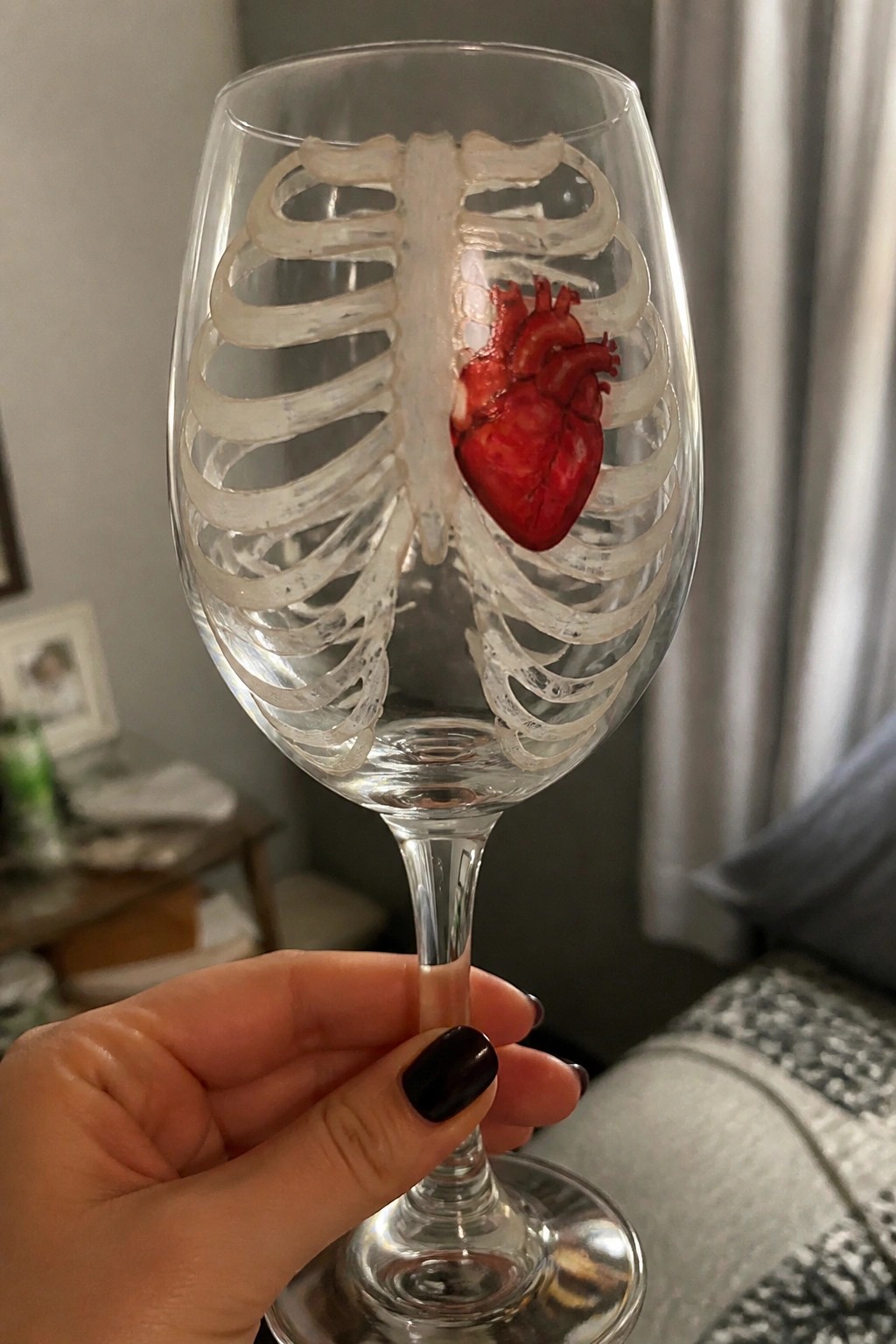 Hand with dark nails holds wine glass with ribcage and red heart.