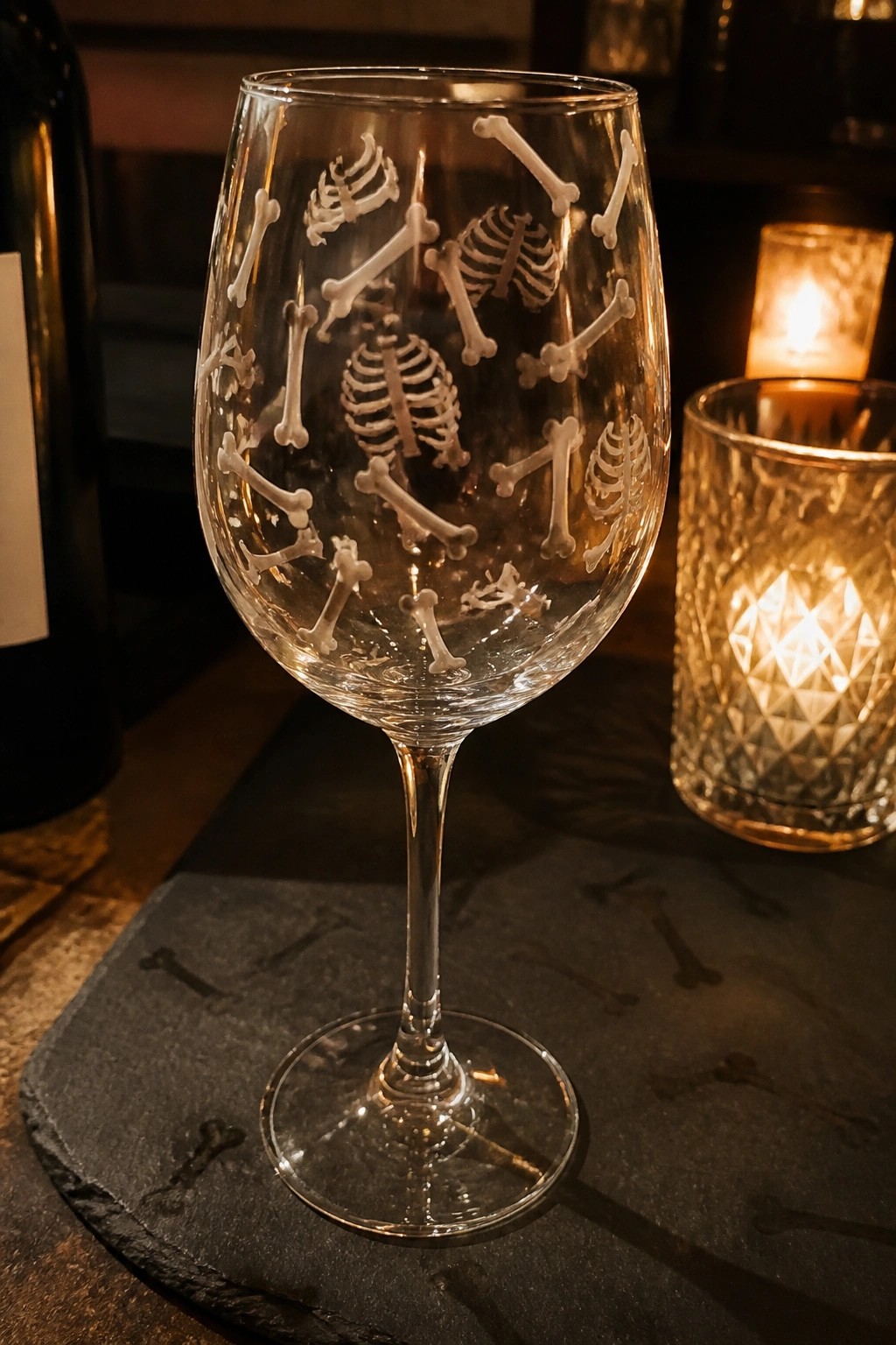 Etched skeleton wine glass on bone-patterned slate beside lit crystal candle holder