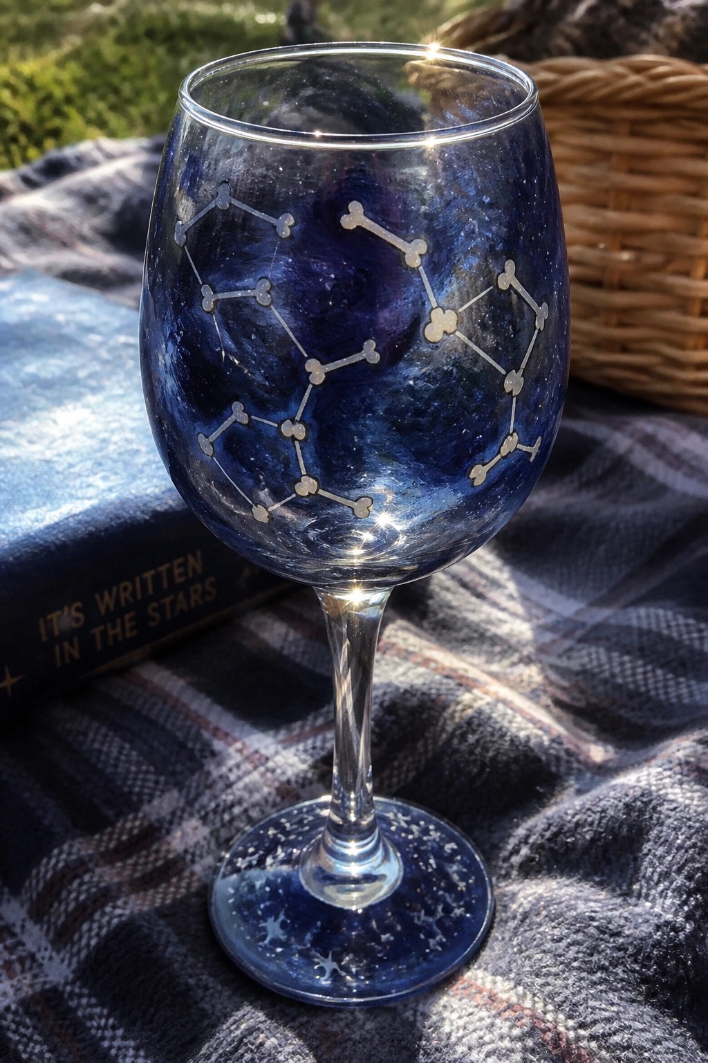 Cosmic blue wine glass with silver constellations beside astrology book on plaid blanket