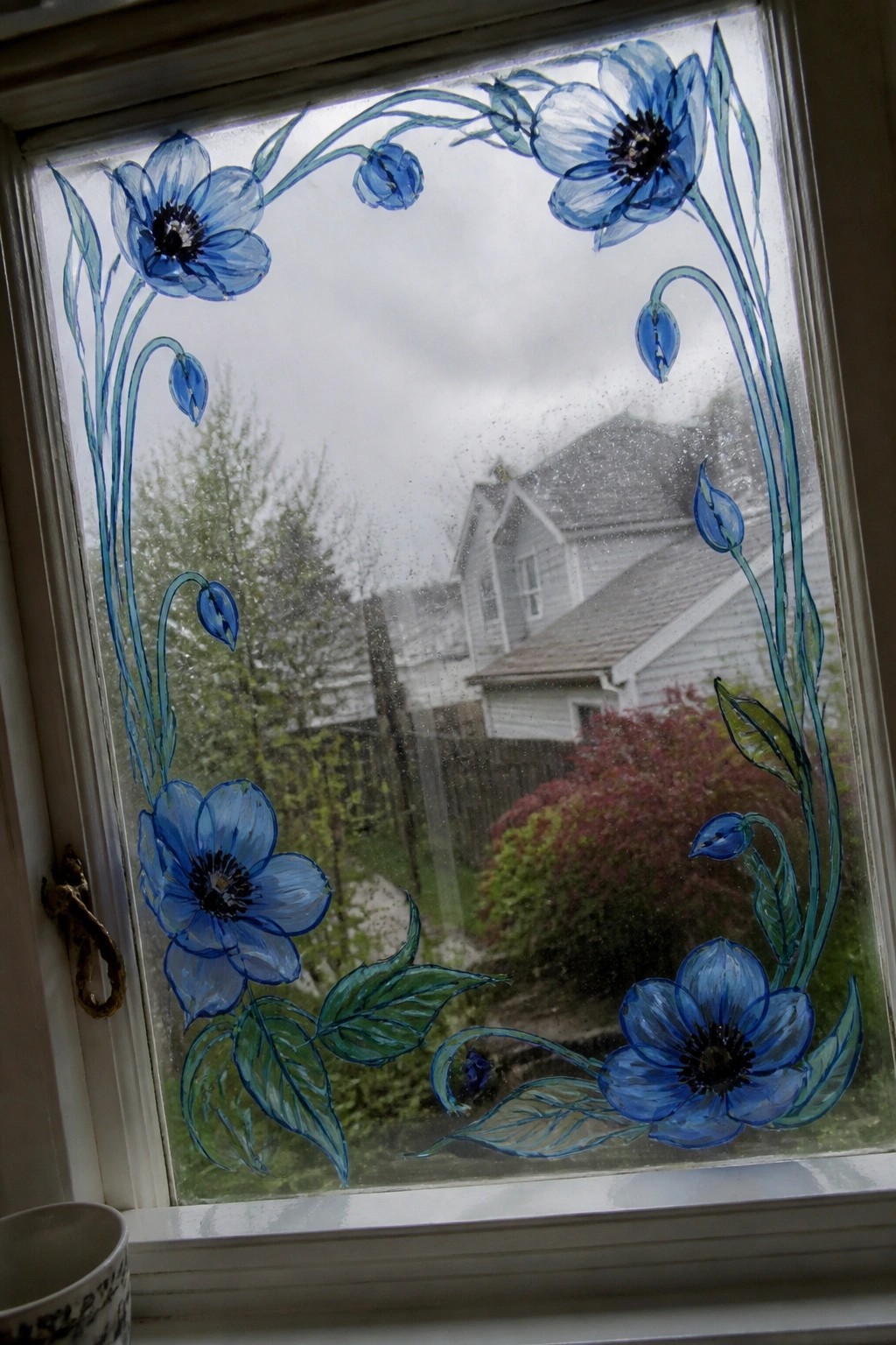 Window with blue painted flowers framing view of houses and trees.